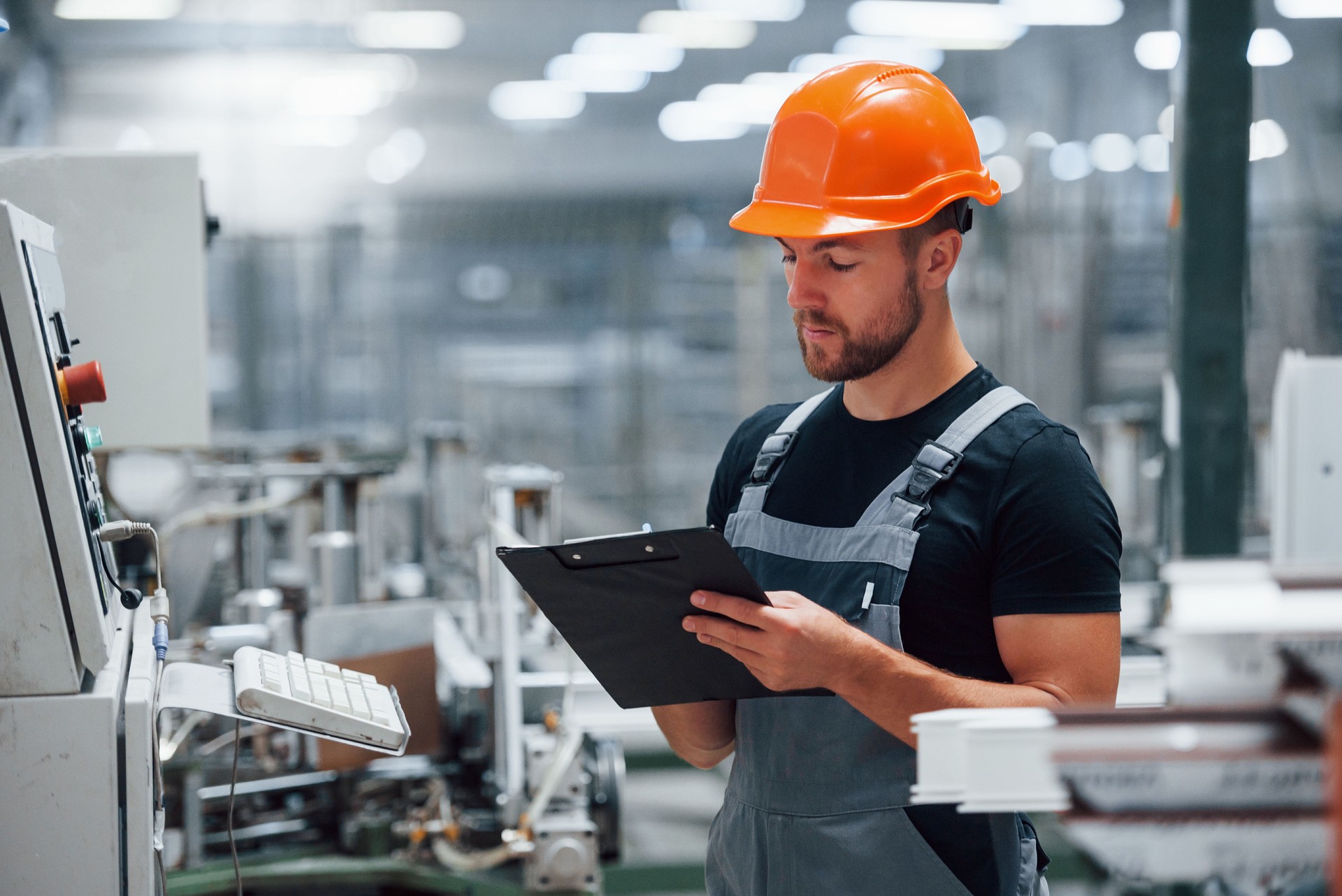Looks at equipment and makes notes. Industrial worker indoors in factory. Young technician with orange hard hat